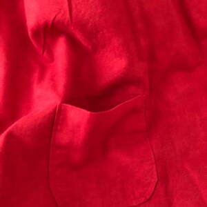 Peut inclure: Un v&ecirc;tement en tissu rouge vif dot&eacute; d'une poche plaqu&eacute;e cousue sur le devant. Le mat&eacute;riau pr&eacute;sente une texture mate et douce avec des plis subtils sur toute la surface, mettant en &eacute;vidence la construction simple de cette pi&egrave;ce textile.