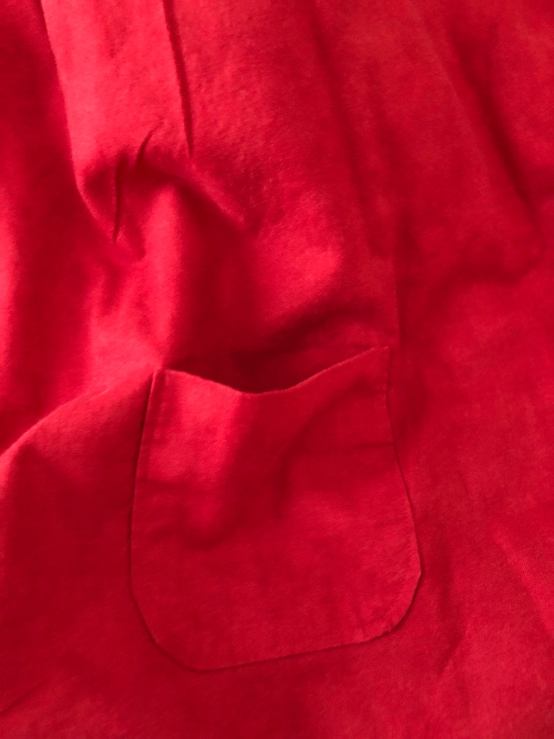 Peut inclure: Un v&ecirc;tement en tissu rouge vif dot&eacute; d'une poche plaqu&eacute;e cousue sur le devant. Le mat&eacute;riau pr&eacute;sente une texture mate et douce avec des plis subtils sur toute la surface, mettant en &eacute;vidence la construction simple de cette pi&egrave;ce textile.