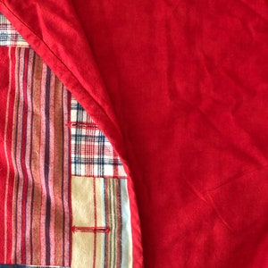 Peut inclure: Un article textile pr&eacute;sentant un panneau de tissu rouge uni repli&eacute; pour r&eacute;v&eacute;ler un envers en patchwork. La section patchwork affiche divers motifs, notamment des rayures verticales rouges, brunes et bleues, ainsi qu'un motif &eacute;cossais.