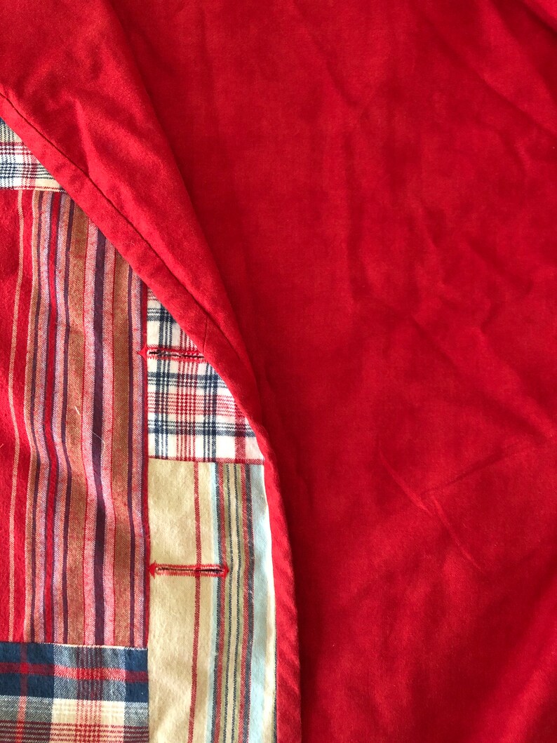 Peut inclure: Un article textile pr&eacute;sentant un panneau de tissu rouge uni repli&eacute; pour r&eacute;v&eacute;ler un envers en patchwork. La section patchwork affiche divers motifs, notamment des rayures verticales rouges, brunes et bleues, ainsi qu'un motif &eacute;cossais.