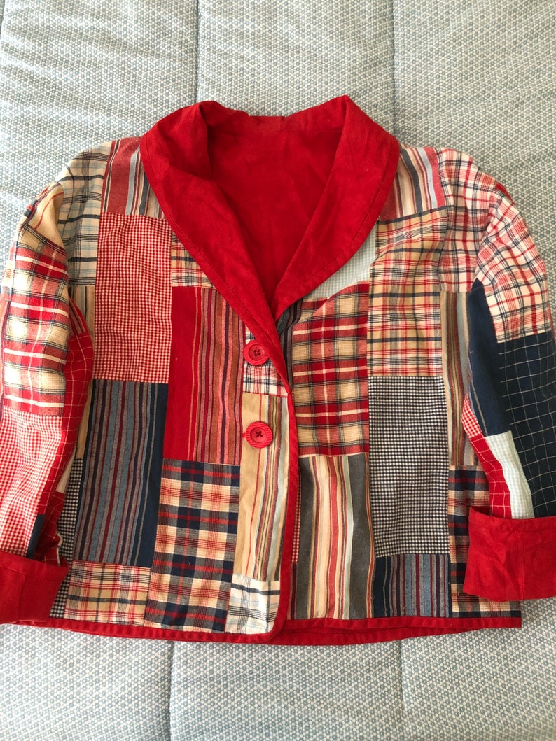 Peut inclure: Une veste en patchwork compos&eacute;e de divers carr&eacute;s de tissu &agrave; carreaux et &eacute;cossais dans des tons rouge, bleu, beige et blanc. La veste pr&eacute;sente un col ch&acirc;le rouge uni, des poignets rouges et deux boutons rouges sur la fermeture avant &agrave; manches longues.