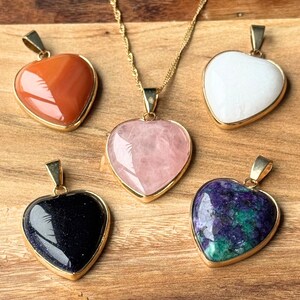 Könnte beinhalten: Fünf herzförmige Anhänger mit goldfarbenen Metallösen. Ein Anhänger ist an einer goldfarbenen Kette befestigt. Die Steine umfassen einen glatten orangen, einen durchscheinenden weißen, einen blassrosa, einen dunkelblauen mit Glitzer und einen violett-grün gesprenkelten Stein.