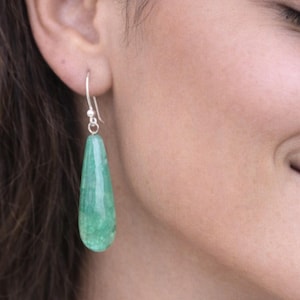 Peut inclure: Gros plan d'une oreille portant une seule boucle d'oreille en forme de goutte d'eau, en pierre verte. La boucle d'oreille est suspendue à un crochet argenté. La pierre a une surface texturée et un léger dégradé de couleur.