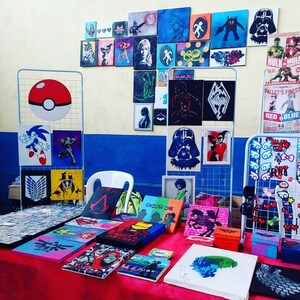 May include: A table display of various hand-painted canvas art featuring popular characters and designs. The art includes characters from video games, anime, and comics. The table is covered in a red tablecloth.