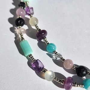 May include: A close-up of a colourful beaded necklace. The necklace features a variety of gemstone beads in shades of purple, pink, turquoise, and white, interspersed with silver-coloured metal beads. The beads are strung together on a delicate chain.