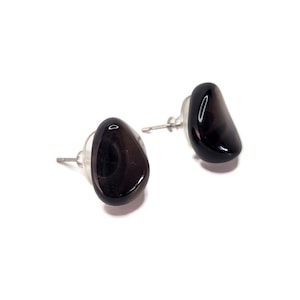 May include: A pair of dark gray, polished stone stud earrings. Each earring has a rounded, irregular shape and is mounted on a silver post with a clear backing. The earrings are set against a white background.