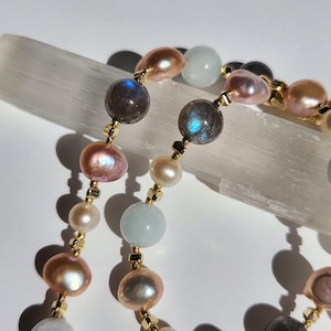 May include: A beaded necklace featuring alternating round iridescent pink pearls, smooth pale blue beads, and dark grey stones with blue flashes. The beads are separated by small gold-toned metallic spacers, creating a delicate and colorful pattern.