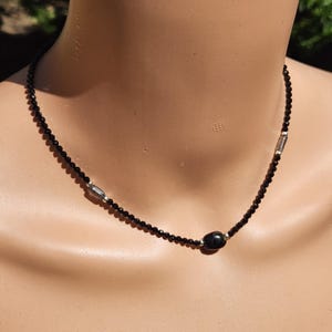 Op de afbeelding: Een ketting met kleine, gefacetteerde zwarte kralen in een doorlopende streng. Het ontwerp bevat twee langwerpige zilverkleurige metalen kralen die symmetrisch zijn geplaatst en één grotere, gladde, ronde zwarte kraal in het midden.