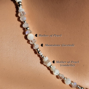 May include: A white necklace with a mix of round and faceted beads. The beads are made of mother of pearl and moonstone. The necklace is strung on a silver chain.