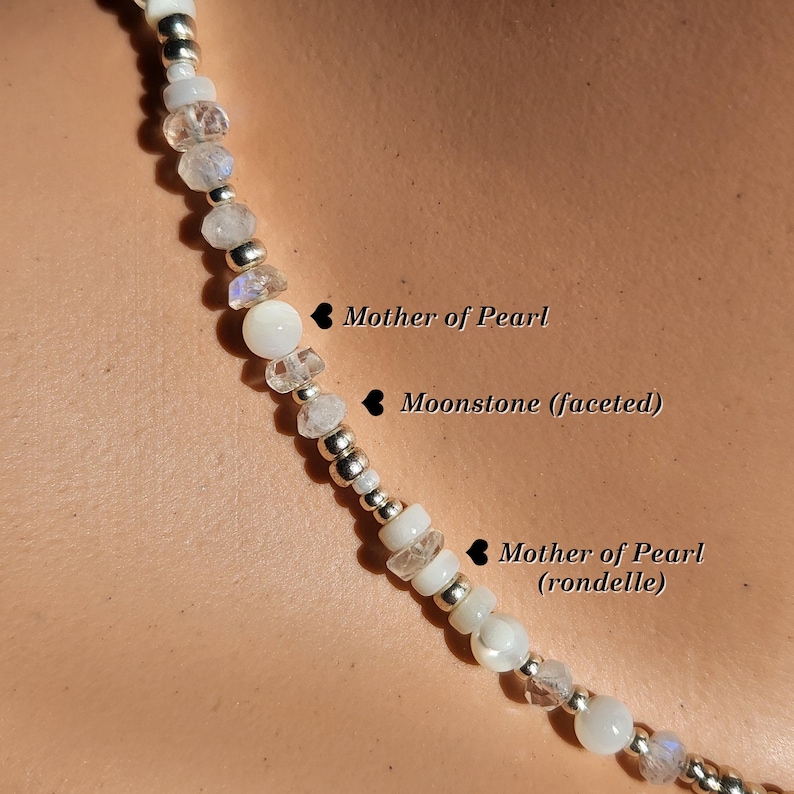 May include: A white necklace with a mix of round and faceted beads. The beads are made of mother of pearl and moonstone. The necklace is strung on a silver chain.