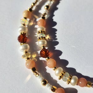 Peut inclure: Gros plan d'un collier multi-rangs de perles avec un mélange de perles blanches, pêche et brunes. Le collier présente de petites perles dorées et des perles plus grandes de formes et de tailles variées.