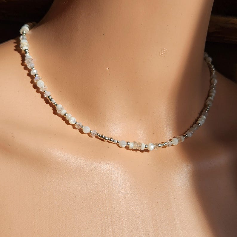 May include: A delicate silver and white beaded necklace with iridescent beads. The necklace is on a white mannequin.