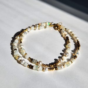 May include: A delicate, layered bracelet featuring a mix of white pearls, iridescent beads, and small gold-colored beads. The bracelet is designed with a gold-colored clasp and is displayed on a white surface, with sunlight casting shadows.