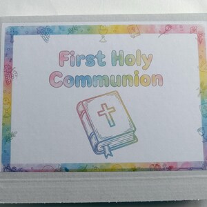 May include: A white cardboard box with a colorful border and the words "First Holy Communion" in pastel colors. The box features an illustration of a Bible with a cross and decorative elements.