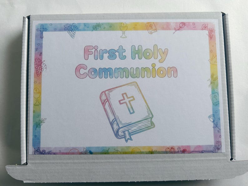 May include: A white cardboard box with a colorful border and the words "First Holy Communion" in pastel colors. The box features an illustration of a Bible with a cross and decorative elements.