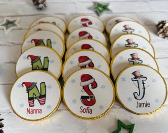 May include: Assortment of round chocolate coins wrapped in gold foil with festive Christmas designs. Each coin features a different letter of the alphabet, a Christmas character, and a name. The names include Nanna, Sofia, and Jamie.