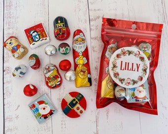 Puede incluir: Una colección de chocolates con temática navideña que incluye figuras envueltas en papel aluminio de Santa Claus, un cascanueces, un oso y muñecos de nieve, junto con barras de chocolate cuadradas y una bolsa roja con el nombre LILLY.