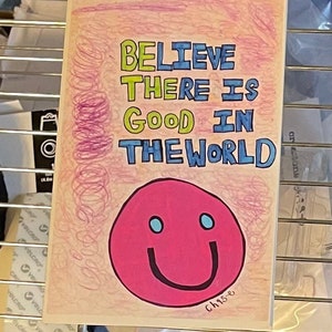 May include: A colorful drawing with the words "BELIEVE THERE IS GOOD IN THE WORLD" in blue, green, and yellow. A pink smiley face with blue eyes and a black smile is at the bottom. The artist's name, "Chase", is in the bottom right corner.