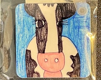 May include: A square keychain featuring a hand-drawn cow face with black and white markings, a pink nose, and blue background. The keychain is attached to a silver key ring and is packaged in a clear plastic bag.