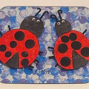 Pode incluir: Uma base para copos retangular com duas joaninhas de desenho animado. As joaninhas têm asas vermelhas com manchas pretas e cabeças pretas. O fundo é um padrão de bolhas azuis e brancas. O nome "ChiSe" está escrito no canto inferior direito.