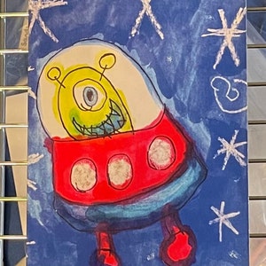 May include: A child's drawing of an alien in a red spaceship against a blue background with white stars. The alien is yellow with one eye and a toothy grin. The name "Chase" is written in the bottom right corner.