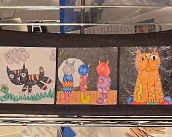 May include: Black pencil case featuring three panels with colorful cat illustrations. The first panel shows a black cat with orange stripes. The second panel depicts three cat-like figures. The third panel shows an orange cat.