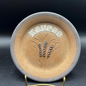 May include: A round, tan-colored ceramic plate with a dark gray rim. The word KANSAS is embossed in white lettering at the top. Below the text, a stylized wheat stalk design is carved into the center of the plate with dark brown accents.