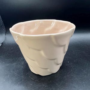 May include: A pale pink ceramic planter featuring a repeating embossed wave or shell-like pattern across its exterior surface. The bowl-shaped vessel has a wide, open top with a slightly scalloped rim and a smooth, glossy finish throughout.
