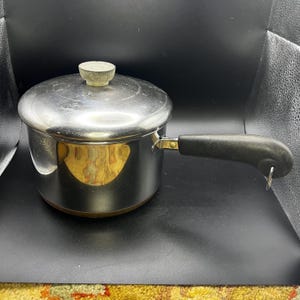 Könnte beinhalten: Ein Edelstahl-Kochtopf mit Deckel und schwarzem Griff. Der Deckel hat einen hellfarbigen Knopf. Der Kochtopf steht auf einer schwarzen Oberfläche.