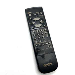 May include: A black Quasar VCR remote control with numerous gray buttons. The remote has buttons for power, play, pause, rewind, fast forward, and channel selection. The brand name "Quasar" is printed at the bottom.