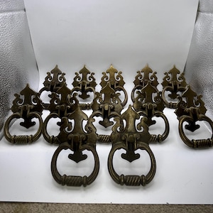 May include: A collection of antique brass drawer pulls. Each pull features an ornate, decorative backplate with a ring handle. The pulls have a vintage aesthetic, with a warm, aged patina. Ideal for furniture restoration or adding a classic touch.