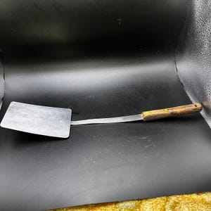 Könnte beinhalten: Ein Edelstahl-Pfannenwender mit rechteckiger Klinge und langem, dünnem Griff. Der Griff ist aus Holz mit dunkelbraunem Finish und gelbem Akzent. Der Pfannenwender ist für das Kochen und die Zubereitung von Speisen konzipiert.
