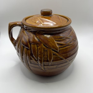 Puede incluir: Una olla de cerámica marrón con tapa y asa. La olla tiene un acabado brillante y está decorada con un patrón de hojas. La tapa tiene un pequeño pomo en la parte superior. La olla probablemente se utiliza para cocinar o servir alimentos.