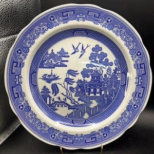 May include: A round ceramic plate featuring a blue and white willow pattern. The design includes a central scene with a pagoda, a bridge, a boat, and two flying birds, surrounded by a wide border with geometric and floral motifs.