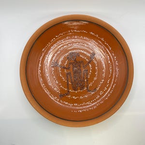 Puede incluir: Un plato de cerámica de color terracota con un borde marrón oscuro y un diseño central. El diseño presenta una figura estilizada dentro de círculos concéntricos, con acentos de puntos blancos. El plato tiene un aspecto rústico y hecho a mano.