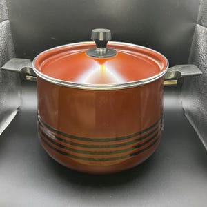 Peut inclure: Une marmite marron avec un couvercle et deux poignées latérales. La marmite a des rayures noires autour de la base et un bouton noir sur le couvercle. L'intérieur de la marmite et du couvercle est d'une nuance de marron plus claire.