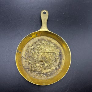 May include: A small, brass-coloured decorative pan with a handle. The pan's interior features an embossed scene of a building, a horse-drawn carriage, and text that reads "Pennsylvania Dutch Country, Lancaster, Penna."