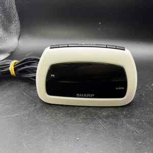 May include: A cream-colored Sharp digital alarm clock with a black display screen. The front panel features the text SHARP, PM, and ALARM. A black power cord is attached to the side, secured with a yellow twist tie. The top has black control buttons.