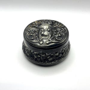 Könnte beinhalten: Kleine, runde, antike Schmuckdose in Silberoptik. Der Deckel und die Seiten sind mit detaillierten Blumen- und Rankenmustern verziert. Die Dose hat ein leicht angelaufenes Aussehen, was ihren Vintage-Charme unterstreicht.