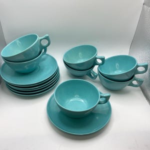 May include: A set of turquoise ceramic teacups and saucers. The cups have a unique handle design and are stacked on top of the saucers. The set includes multiple cups and saucers, perfect for serving tea or coffee. The color is a vibrant, solid turquoise.