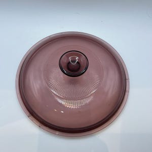 Puede incluir: Una tapa redonda de vidrio morado transparente con un pomo central. La tapa presenta un diseño acanalado que irradia desde el pomo. El pomo tiene un pequeño asa circular. La tapa está diseñada para utensilios de cocina.