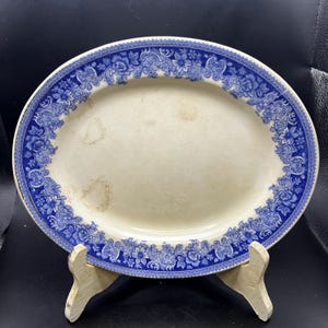 Puede incluir: Plato ovalado para servir con un centro color crema y un borde floral azul. El borde azul presenta intrincados diseños florales y de volutas. El plato se exhibe sobre un soporte de madera, mostrando su estética vintage.