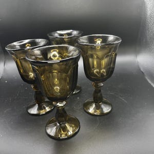 May include: Set of four vintage-style, olive-green stemmed glasses. Each glass features a decorative, fluted bowl, a detailed stem, and a round base. The glasses are arranged on a dark surface, showcasing their elegant design.