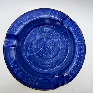 May include: A round, cobalt blue ashtray with a brown rim. The ashtray features a white etched design of two fish and the words "FISCHERS TUB" and "WEESEN RESTAURANT". The ashtray has three grooves for resting cigarettes.
