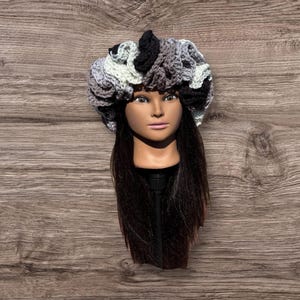 May include: A handmade crochet hat in a variety of colors, including black, gray, and white. The hat is designed with a textured, open weave pattern. The hat is displayed on a mannequin head with long, dark hair.