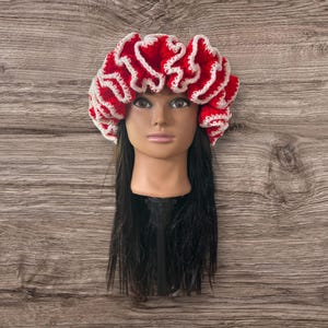 May include: A red and white crocheted head covering with a ruffled design. The head covering is displayed on a mannequin head with long black hair. The background is a wooden surface.