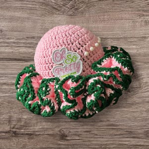May include: Pink crocheted sun hat with a green ruffle trim and a pink and green embroidered patch that says "Oh So Pretty".