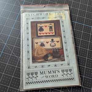 May include: A clear plastic bag containing a craft pattern titled "Patchwork Pals" and "Mumm's the Word." The pattern features folk art style images of figures, hearts, and a basket. The background is a light blue color.