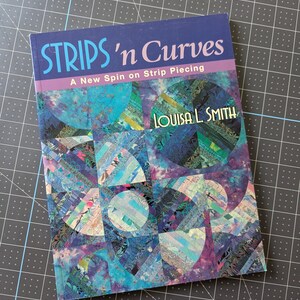 Op de afbeelding: Een instructieboek met zachte kaft getiteld Strips 'n Curves: A New Spin on Strip Piecing door Louisa L. Smith. De kaft toont een levendig quiltontwerp bestaande uit blauwe, paarse en groene stoffen stroken in cirkelvormige patronen.