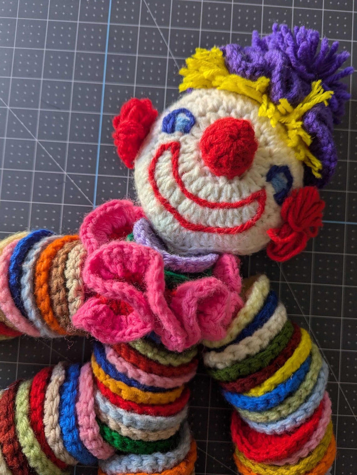 May include: A crocheted clown doll with a white face, red nose, and blue eyes. The doll has a pink ruffle around its neck and is wearing a multi-colored striped outfit.